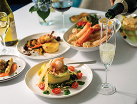 A bubbly glass of champagne being poured in front of a variety of gourmet plates on a white tablecloth, served on American Cruise Lines.