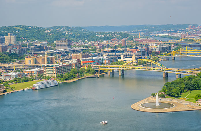 An aerial view of the Ohio River, containing a Pittsburgh cityscape with mountains in the distance, several yellow bridges over the river, and American Heritage paddlewheeler docked along the river. 