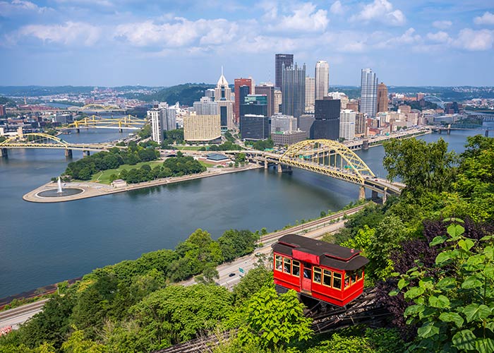 A red cable car descends a forest hill overlooking the Ohio River and a Pittsburgh cityscape featuring a brideg with a yellow truss in the middle and  several skyscrapers under a partly cloudy sky.