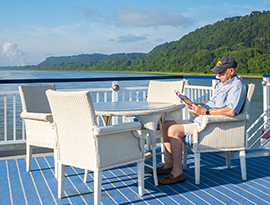 A man sits on a white wicker chair at a round white table on an American Riverboat's sun deck while reading a book with views of the Ohio River in the distance.