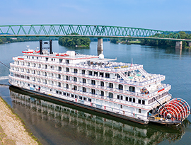 American Heritage paddlewheeler docked letting passengers off from the front of the white ship. It is decorated with patriotic red white and blue bunting flags on every railing. In the background, there is a green bridge above the Ohio River.