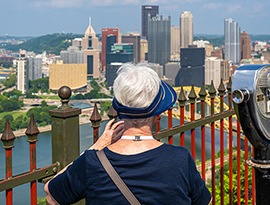 A woman with short white hair is wearing a visor while looking off into the distance at the cityscape and Ohio River behind a red rusted fence. To her right, there is a Tower Viewer.