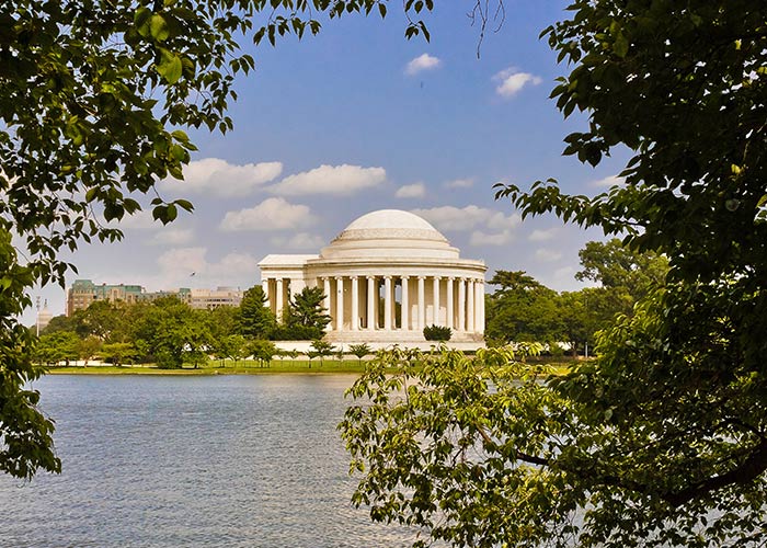 Jefferson Memorial, a round, white building with countless tall pillars surrounding it and a round dome on the roof in Washington, D.C. The waters of the Potomac River and leafy tree branches are in the foreground.