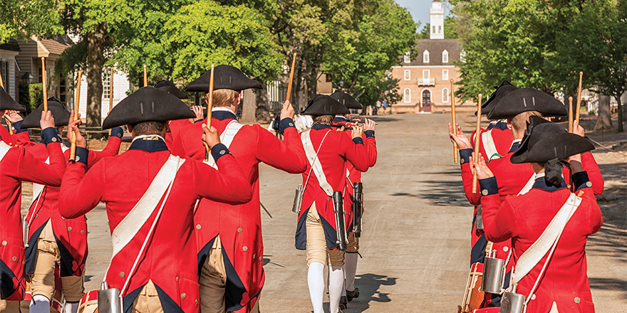 A parade of people in Colonial Williamsburg dressed in colonial costumes with bright red shirts, black hats, white knee-high socks, and khaki pants that stop above their knees. They are holding drumsticks in their hands, and a few people in the front are playing the flute. There are trees on each side and a brick building at the end of the path.