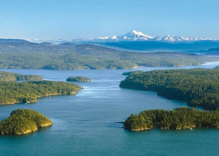 An aerial view of Puget Sound and the various sizes of the San Juan Islands along the water, and the snow-capped Mount Rainer in the distance.