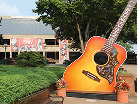 A large guitar statue with "Grand Ole Opry" written along the neck surrounded by tall trees. In the background, the Grand Ole Opry is present with a large red poster with many famous country artists faces on it above the entryway.