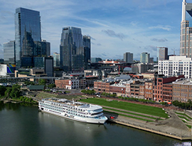 The American Riverboat American Serenade docked on the Cumberland River with an aerial view of Nashville's city landscape, including brick buildings and tall skyscrapers.