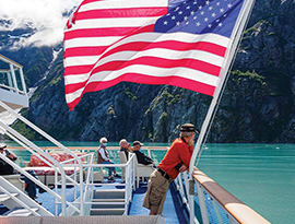 A man leans his arms on the railing of the sun deck on a Constellation Class Coastal Cruise Ship next to a large American flag blowing in the wind while sailing along the Inside Passage. There are several other passengers sitting on the sun deck looking at the mountains that line the waters.