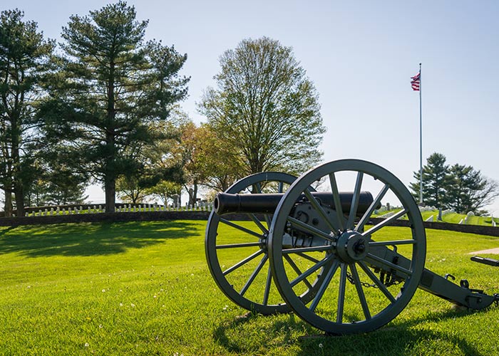 A large black cannon in the middle of Shiloh National Military Park in Savannah, with large trees and an American flag on a tall flag pole in the distance.