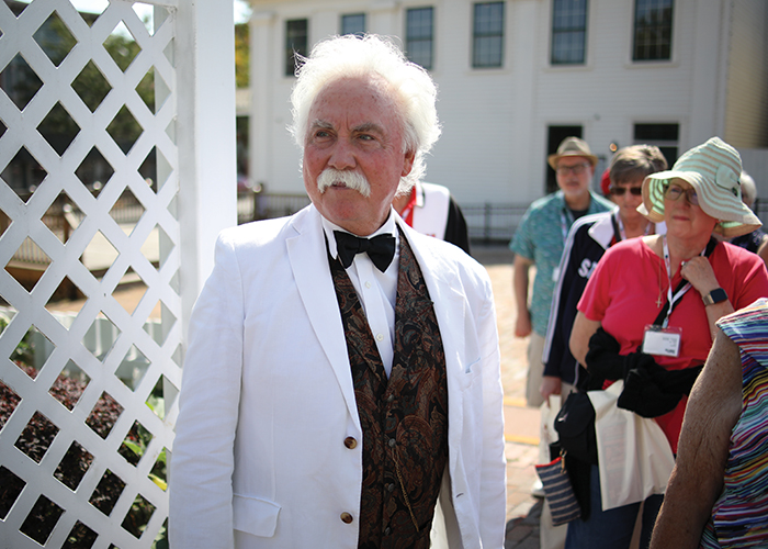 A distinguished older man impersonating Mark Twain with white hair and a white mustache, wearing a white suit and bow tie, showcasing a refined and classy appearance.