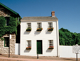 Mark Twain's light cream colored house with a dark green door and window planters to the right of a brown brick giftshop covered with green vines and topped with a green roof surrounded by the whitewashed fence of Tom Sawyer in Hannibal.