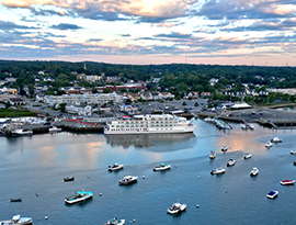 An aerial view of an American Cruise Lines Coastal Cat docked in Plymouth among several small boats on the water. On the shore, there are several buildings and thick trees under a cloudy sunrise.