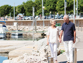 A couple holding hands and smiling at each other while walking along a sidewalk next to the harbor where boats are docked.