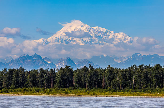 Thick forests line the waters of Cook Inlet in Anchorage. Small mountains peak over the trees, and the snow-capped Mount Denali is in the distance, tall enough that it is surrounded by the clouds.