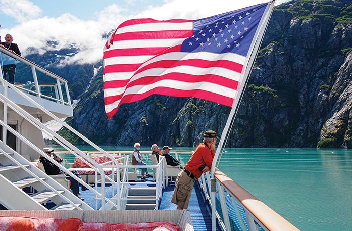 A man leans his arms on the railing of the sun deck on a Constellation Class Coastal Cruise Ship next to a large American flag blowing in the wind while sailing along the Inside Passage. There are several other passengers sitting on the sun deck looking at the mountains that line the waters.