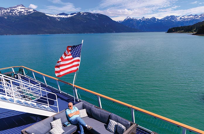 Aerial view of the a man sitting on a L-shaped couch with grey and white pillows on the sun deck on a Constellation Class Coastal Ship that is sailing through the Inside Passage. There is an American flag on a pole connected to the railing that is blowing in the wind. Snow-capped mountains line the water.