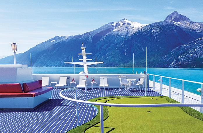 The sun deck of a Constellation Class Coastal Cruise Ship sailing on the crystal blue waters of the Wrangell Narrows with snow-capped mountains in the near distance. The sun deck has a perfectly maintained putting green, red benches, white lounge chairs, and a small round table surrounded by chairs.