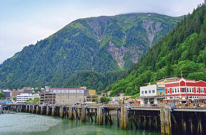 Buildings stand upon a foundation on tall stilts above the Gastineau Channel in Juneau, where a large mountain covered in thick forest is in the near distance.