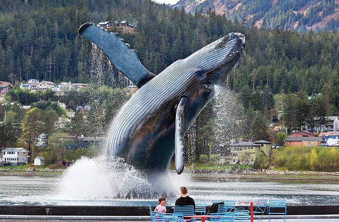 Tahku, a bronze, life-size breaching humpback whale sculpture at the Juneau Whale Fountain. It is rising from an infinity pool in front of the Gastineau Channel located in Juneau. A woman and a child sit on blue chairs surrounding red tables in front of the statue. There are trees and houses in the background. 