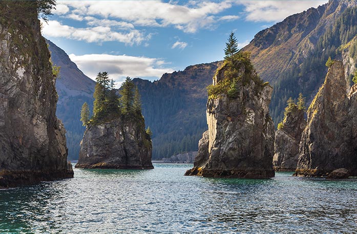 Mountains stand tall around the waters of Kenai Fjords National Park. There are a few tall and narrow  mountain-like structures that are surrounded by the water.