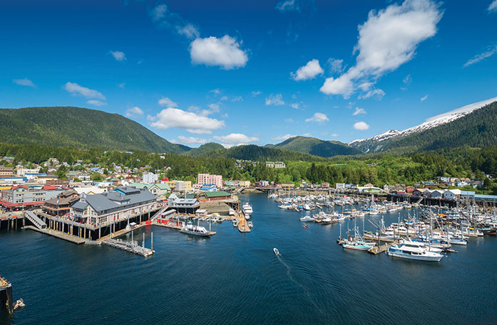 An aerial view of the town of Ketchikan, with several boats docked on the water and the green mountains of Tongass National Forest in the background. 