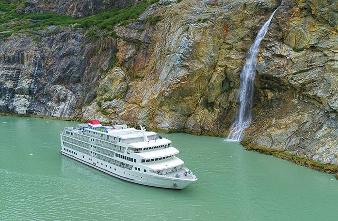 The American Constellation Coastal Cruise Ship sailing through the light green colored passage of Tracy Arm Fjord, surrounded by a cliff with a narrow waterfall.