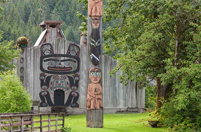 A totem with its art depicting a person sitting down below a black fish with a human face on its tail. Behind the totem, there is a wood fence with a black, red, and blue painting of an animal. The structures are surrounded by greenery.