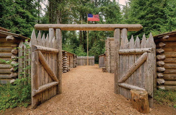 Fort Clatsop, a historic camp with a wooden gate opening to several other wooden structures in Astoria. There is a flag pole with an American flag flying at the top of it, with tall trees in the background.