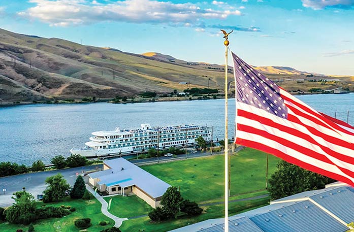 An American Riverboat docked on the Columbia River Gorge along the shores of Clarkston, where there is a building surrounded by open grassland, and a large American flag flying from a roof in the foreground. 