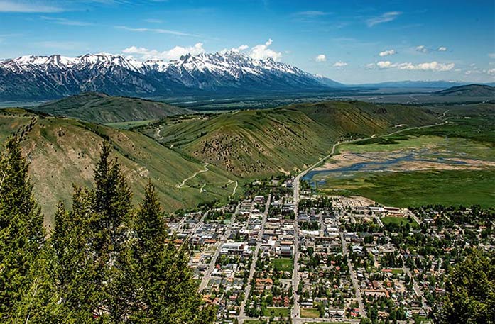 An aerial view of the Jackson cityscape, where buildings cover the ground below tall mountains. In the distance, there are even larger view of the snow-capped Teton Mountain Range.