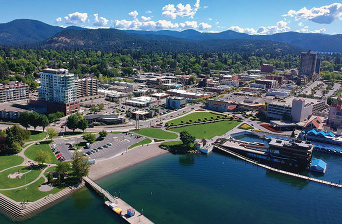 An aerial view of the city and lake of Coeur d’Alene. There are many buildings lining the shoreline, with vast forests and mountains in the distance.