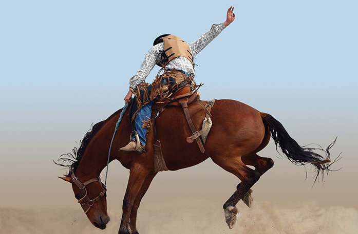 A person riding on a dark brown horse with one hand on the rope and one hand in the air behind him at a rodeo in Pendleton. The horses back feet are in the air, kicking up dirt, and its head is facing the ground.