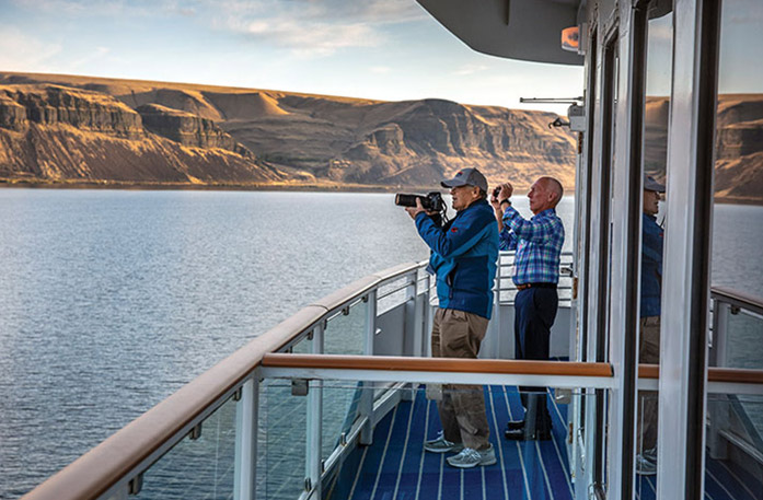 Two American Cruise Lines cruise guests taking photos of the scenic desert canyon mountain views from the deck of an American Riverboat. One of the guests is taking a photo with a large camera, while the other is using his phone camera.