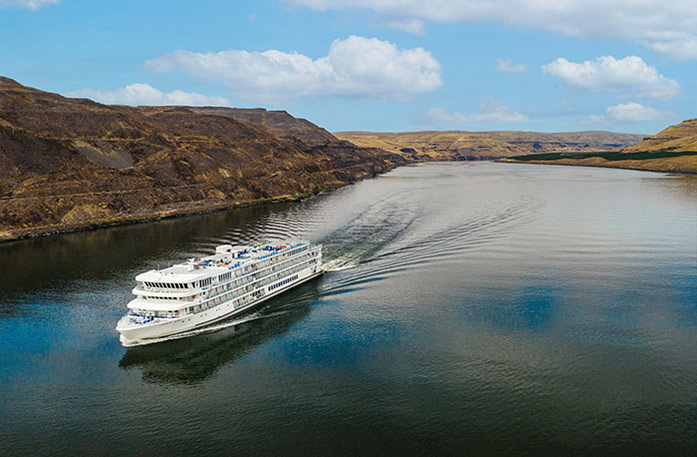 An aerial view of an American Riverboat cruising through the Columbia River Gorge, where there are cliffs alongside the water.