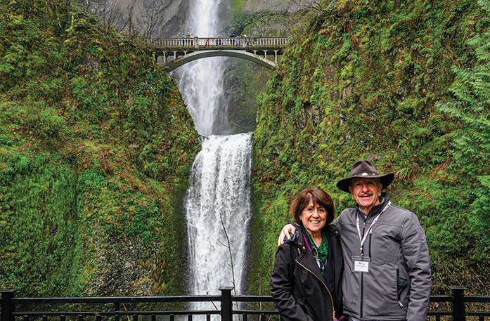 A couple is smiling for a photograph in front of Multnomah Falls, a tall, rushing white waterfall with a bridge connecting two forest-covered mountains. There are several people walking along the bridge.