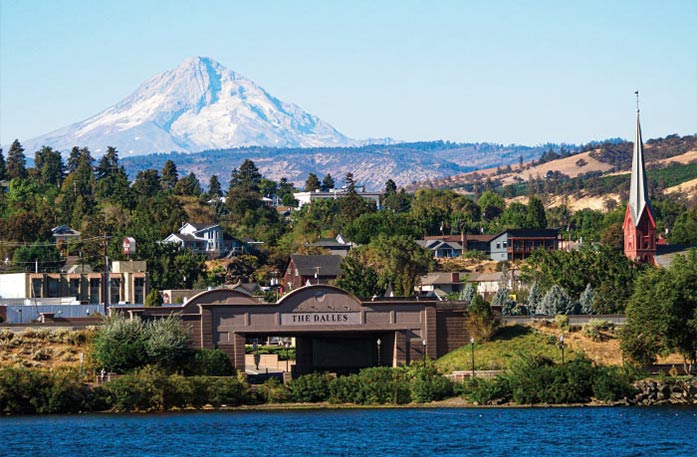 A brown brick tunnel with "The Dalles" written in the middle stands alongside the Columbia River, surrounded by thick greenery and large hills. Mount Hood and Mount Adams meet in the distance in The Dalles.