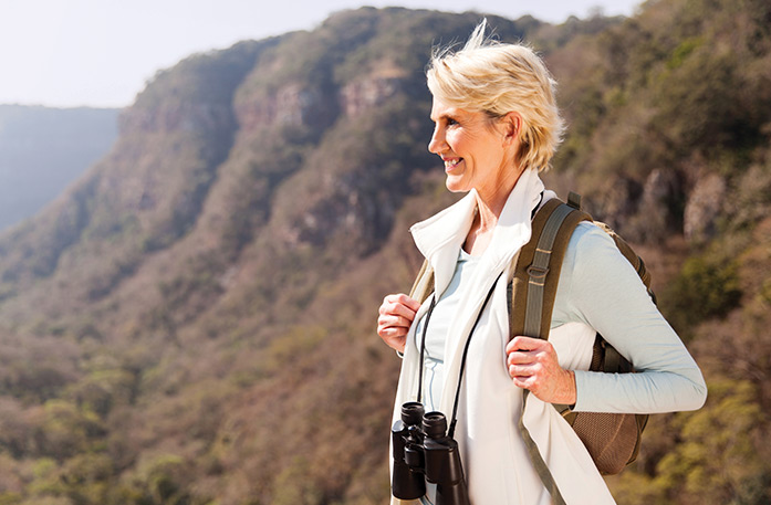 A woman with short blonde hair with binoculars around her neck and a brown backpack on her shoulders is smiling towards the left, with grass-covered cliffs out of focus in the background.