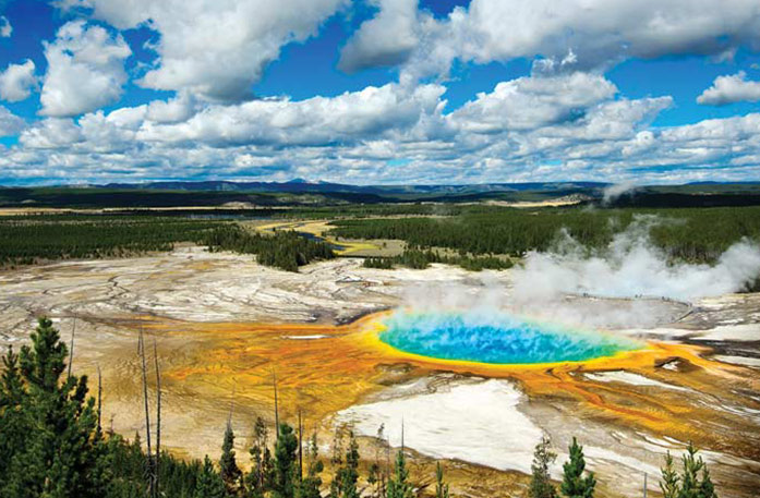 An orange and yellow circular hot spring with large white steam coming off of the bright blue water, surrounded by the forests and mountains of Yellowstone National Park.