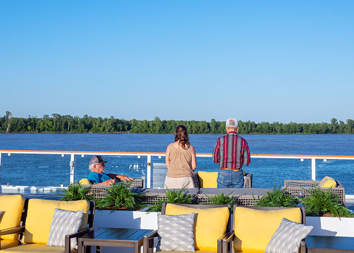 On an American Cruise Lines Coastal Cat Sun Deck, a couple is standing against the glass railing gazing at the Florida coast, the trees lining the horizon line, and the clear blue sky. There are four patio chairs with yellow and grey pillows in the foreground, and in between those and the couple, there is a man sitting on a chair facing the right.