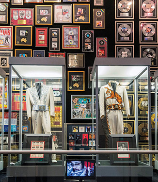 An Elvis Presley exhibit at Graceland in Memphis, with two of his iconic white outfits displayed in glass cases with a wall filled with his records and posters.