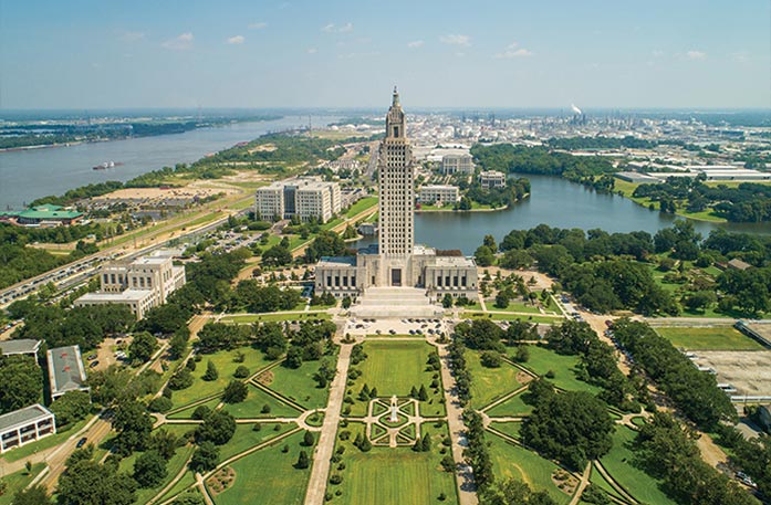 Aerial view of the Louisiana State Capitol building surrounded by landscaped gardens, trees, and a river winding through the Baton Rouge cityscape in the background. 