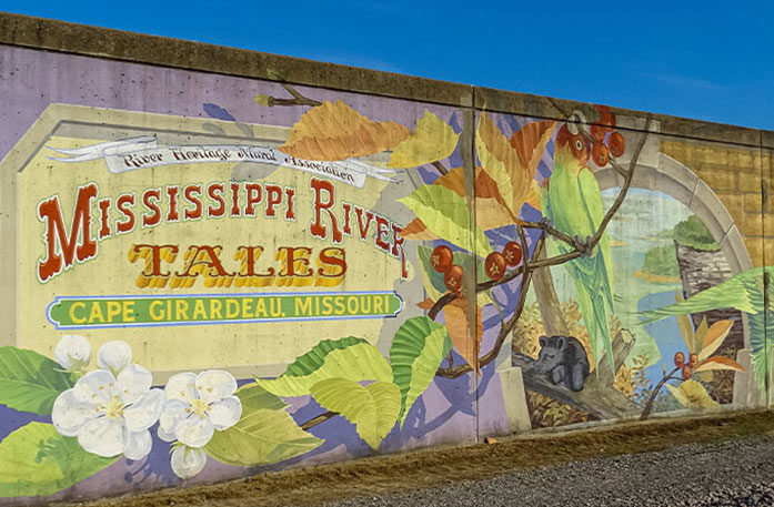 A mural painted on a wall in Cape Girardeau that depicts a parrot sitting on a branch above a small critter on a log surrounded by green leaves and white flowers. To the left, the words "Mississippi River Tales Cape Girardeau, Missouri" are written inside a yellow border on top of a lavender background. "River Heritage Mural Association" is written on a white banner above these words.