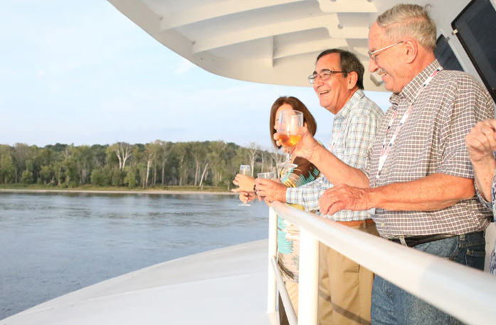 Two men and one woman holding drink glasses and laughing while leaning over the American Riverboat deck railing with the Cumberland River in the background.