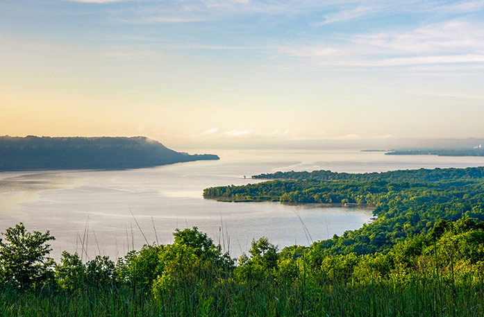 A serene landscape along the Mississippi River surrounded by greenery under a light orange sunrise.