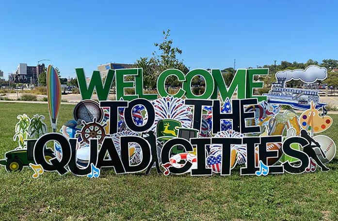 A colorful outdoor sign in Davenport reads "Welcome" in green and  "to the Quad Cities" in black, both with white outline, featuring separated graphics of an art palette, a baseball, a life raft, a tractor, the John Deere green logo with a yellow deer, a steam boat, fireworks, and music notes.