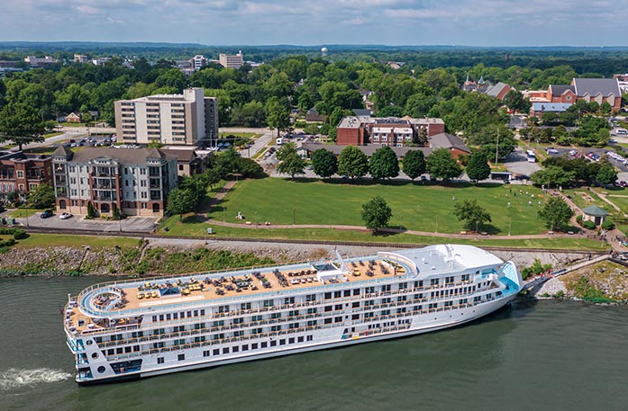 The American Riverboat American Symphony docked along the Tennessee River with its retractable bow gangway letting passengers off the ship in Decatur, with an aerial view of the sun deck on the top of the ship. On land, there is a large empty grass area surrounded by a few buildings and trees.