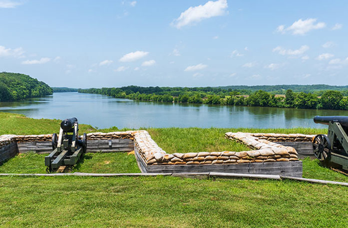 Fort Donelson National Battlefield in Dover, where two cannons are positioned on a grassy fortification overlooking the Mississippi River and lush green landscape under a blue sky.