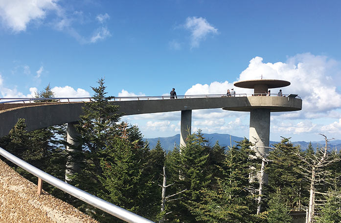 Clingmans Dome, a ramp that curves through tall pine trees leading to a circular observation tower that looks out at the Great Smoky mountains in the distance. There are a few people walking on the ramp and standing on the observation tower.