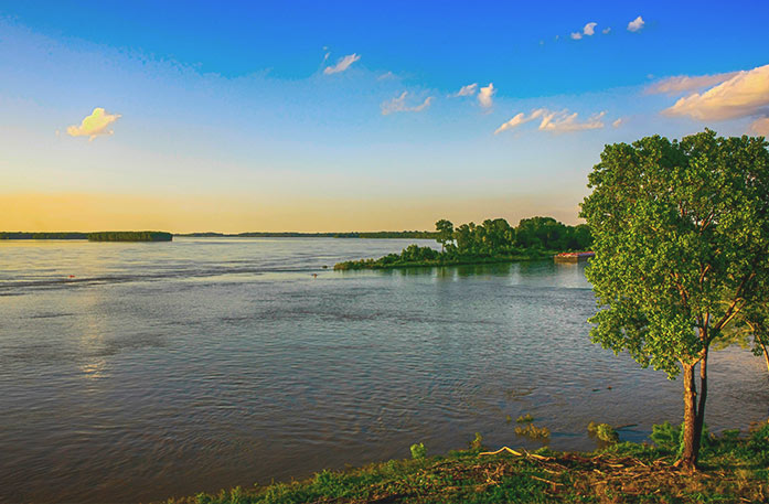 The serene Mississippi River at sunset, featuring calm water, a vibrant blue sky with a hint of the yellow sunset along the horizon, and a green tree in the foreground.