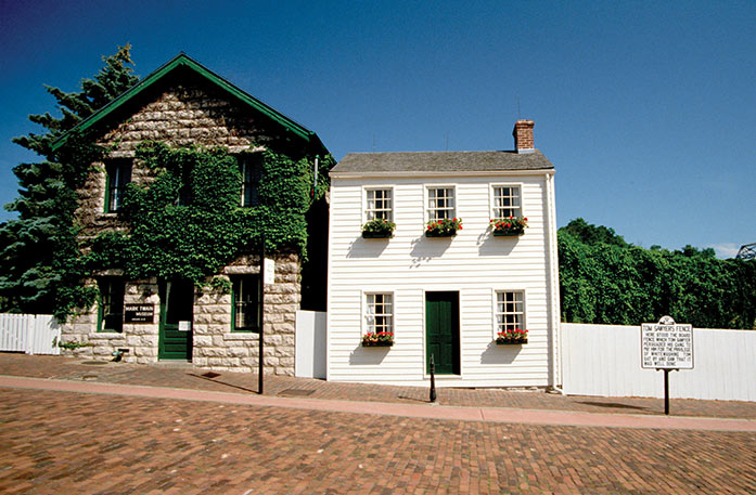 Mark Twain's light cream colored house with a dark green door and window planters in Hannibal to the right of a brown brick giftshop covered with green vines and topped with a green roof surrounded by the whitewashed fence of Tom Sawyer in Hannibal.